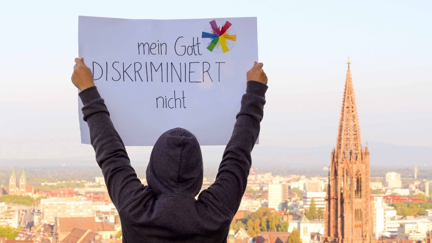 Eine Person hält ein PLakat hoch mit der Aufschrift "Mein Gott* diskriminiert nicht"