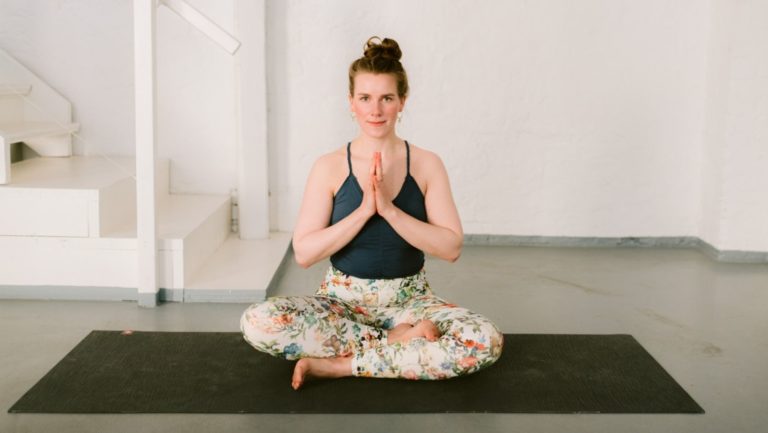 Kathi sitzt im Schneidersitz auf ihrer Yogamatte und hat die Handflächen zusammengelegt